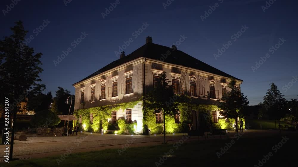 nicely lit old house covered with plants in a garden at night with a few people around nicely lit old house covered with plants in a garden at night with a few people around