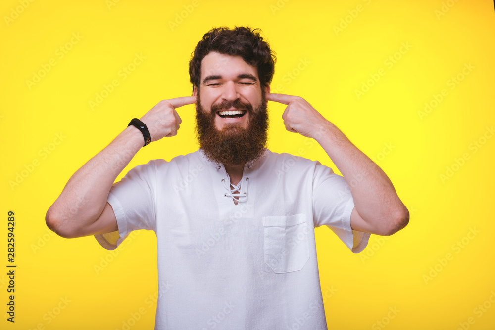 Bearded smiling man is covering his ears with his fingers on yellow backgreound. Bearded smiling man is covering his ears with his fingers on yellow backgreound.