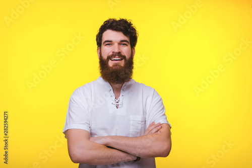 Bearded man is smiling at the camera with crossed arms on yellow backgrounnd.