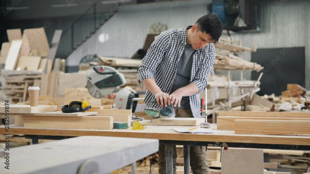 Good-looking carpenter is polishing piece of timber with electric machine in workroom standing at worktable holding equipment. Business and technology concept. Good-looking carpenter is polishing piece of timber with electric machine in workroom standing at worktable holding equipment. Business and technology concept.
