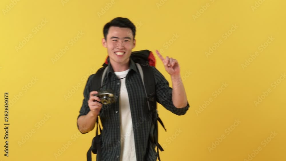 Portrait of happy young Asian tourist man with backpack clicks photo on camera and waves his hand to say goodbye and walks away against yellow background Portrait of happy young Asian tourist man with backpack clicks photo on camera and waves his hand to say goodbye and walks away against yellow background