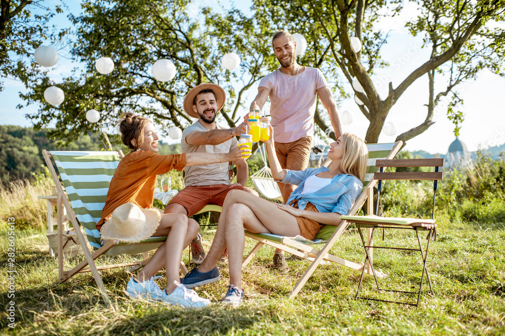 Young and cheerful friends relaxing on the sunbeds, clinking glasses in the beautifully decorated backyard or garden during a festive meeting Young and cheerful friends relaxing on the sunbeds, clinking glasses in the beautifully decorated backyard or garden during a festive meeting