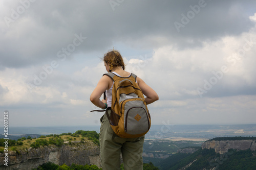 a girl-traveler in a backpack looks into the distance from a cliff on the ruins of a medieval fortified city