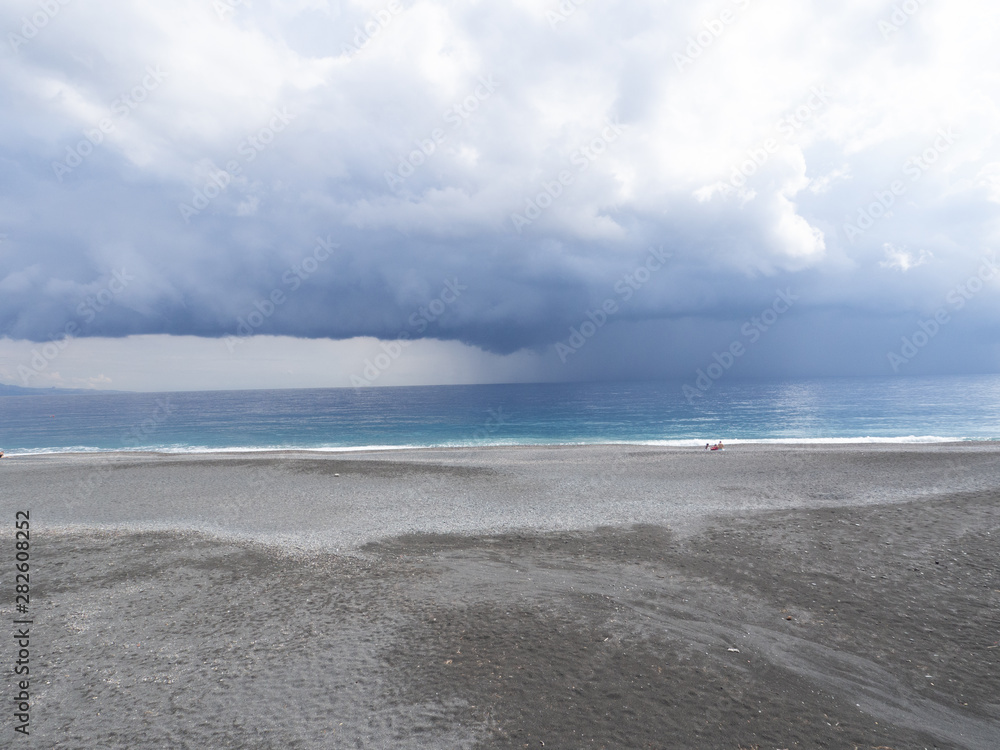 Obraz premium Distant rain and dramatic dark sky over sea with gravel beach in foreground