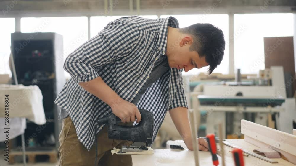 Young carpenter is using jigsaw power instrument to saw wood in workshop manufacturing handmade wooden furniture. People, business and woodwork concept. Young carpenter is using jigsaw power instrument to saw wood in workshop manufacturing handmade wooden furniture. People, business and woodwork concept.