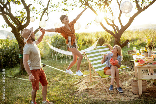 Young friends having fun, walking on a slackline during a picnic in the beautifully decorated garden on a sunset photo