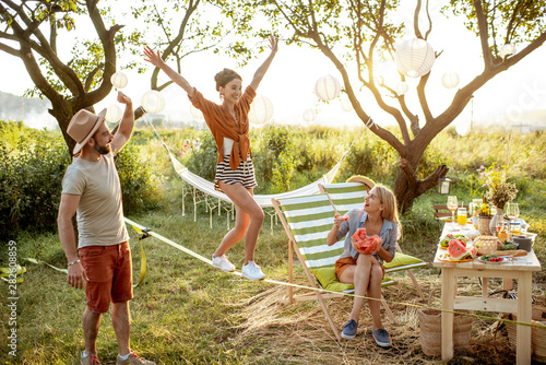 Young friends having fun, walking on a slackline during a picnic in the beautifully decorated garden on a sunset photo