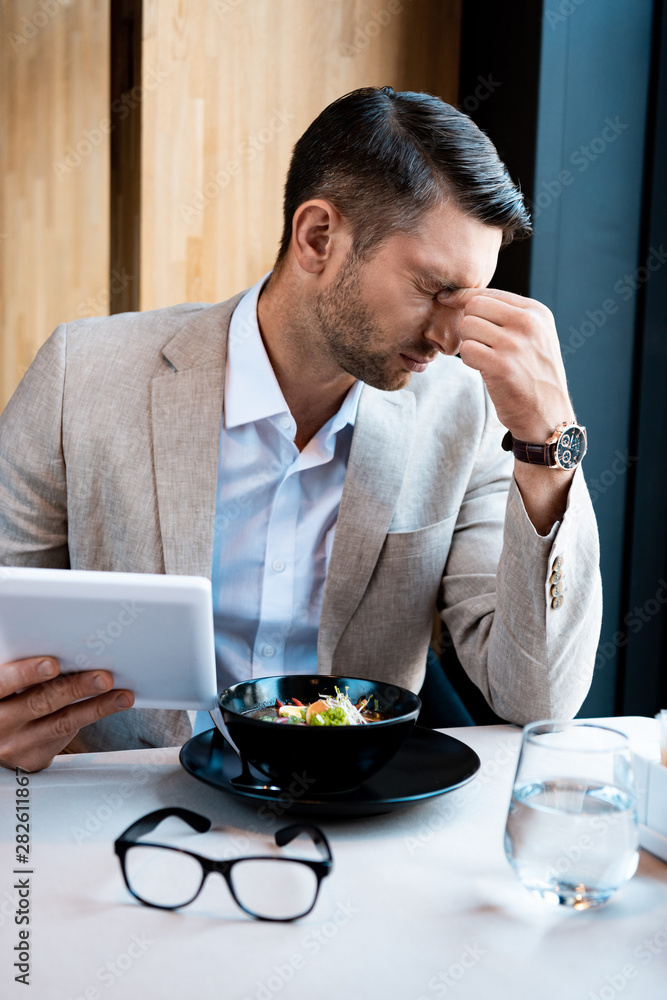 tired businessman touching nose with closed eyes and holding digital tablet in cafe tired businessman touching nose with closed eyes and holding digital tablet in cafe