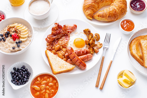 Top view flatlay with varieties of fresh breakfast: fried eggs with bacon and sausages, oatmeal with berries, fried toasts with jam and butter. White background.