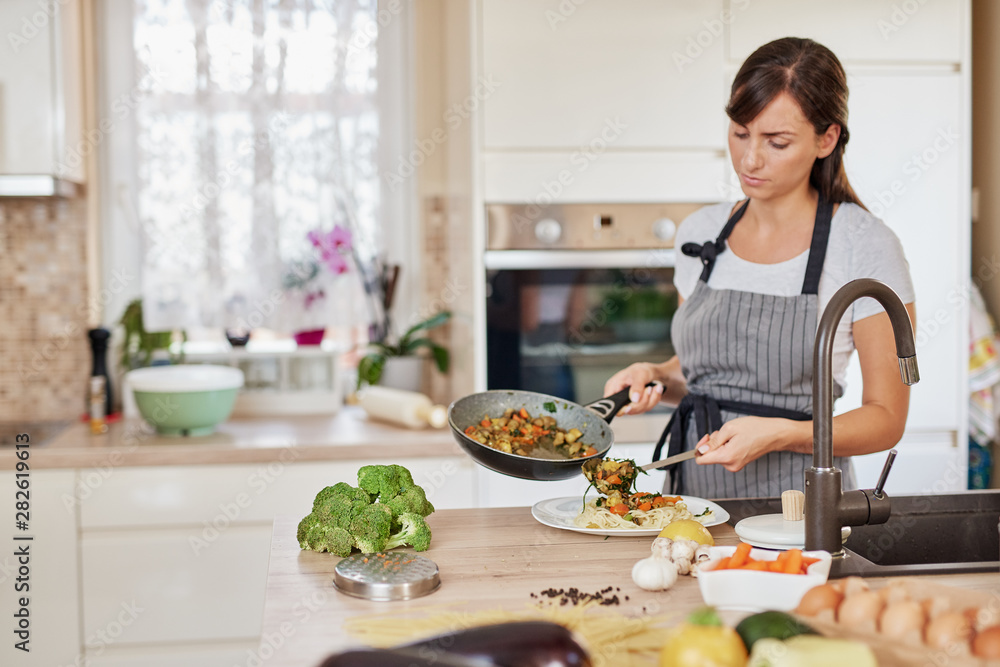 Caucasian woman in apron standing in kitchen and putting sauce on spaghetti.Preparation of italian food concept. Caucasian woman in apron standing in kitchen and putting sauce on spaghetti.Preparation of italian food concept.