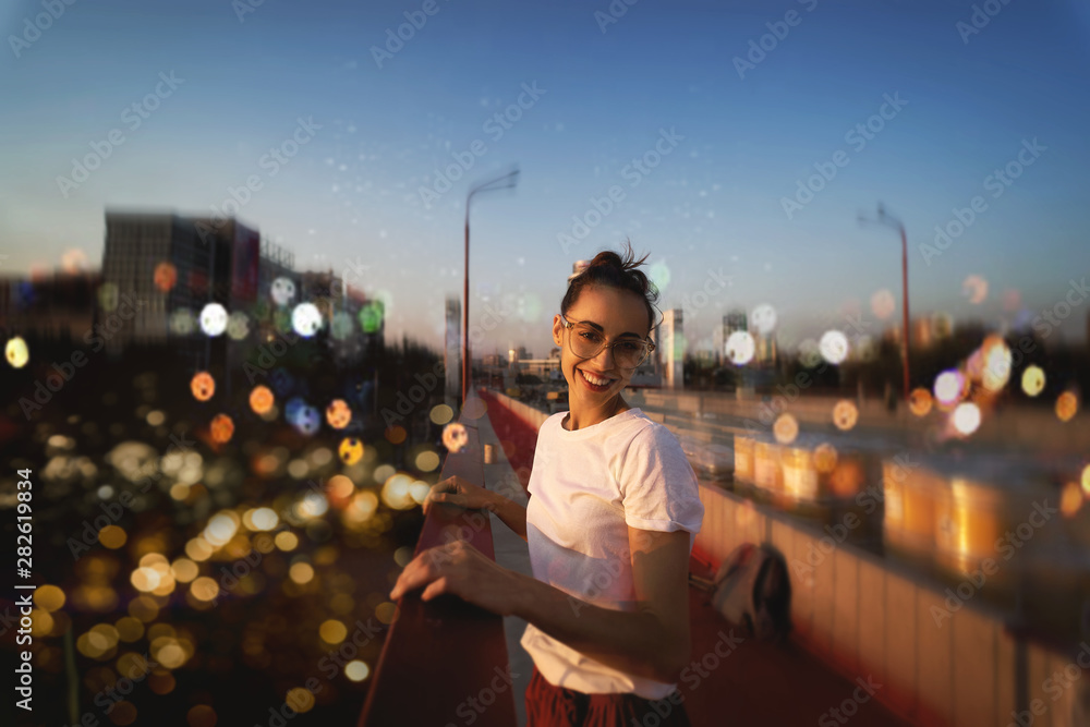 Bright summer lifestyle portrait of young pretty woman in eyewear, red skirt and white T-shirt, standing on the bright red bridge at evening with city background and blurred bokeh. - obrazy, fototapety, plakaty 