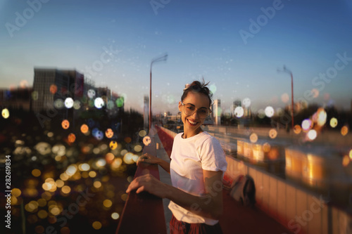 Bright summer lifestyle portrait of young pretty woman in eyewear, red skirt and white T-shirt, standing on the bright red bridge at evening with city background and blurred bokeh. photo