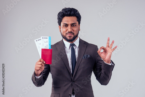 happy indian student male bearded wearing stylish classic suit holding passport airline ticket white studio background . lifestyle summer trip concept