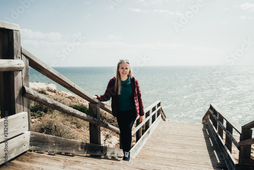 Blond woman standing on stairs near ocean. Attractive girl posing on wooden path. Female enjoying ocean view on the beach in Portugal.
