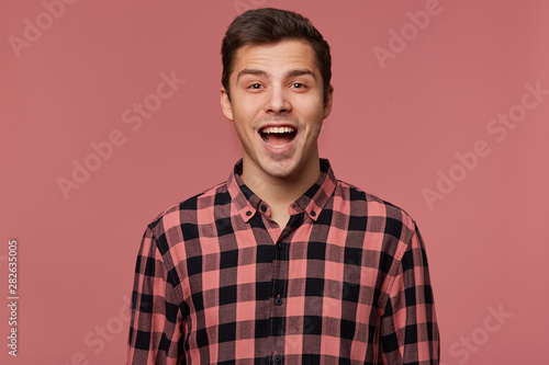 Portrait of young attractive surprised man in checkered shirt, looks at the camera with wide open mouth, stands over pink background.