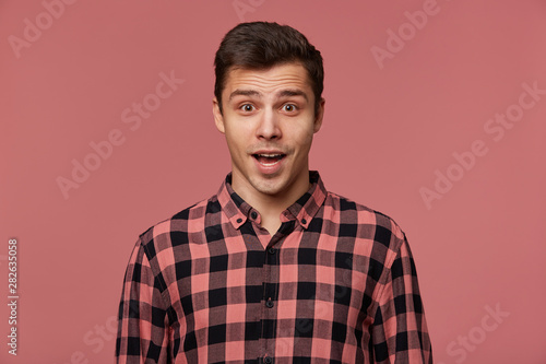 Young handsome wondered guy wears in checkered shirt, looks at the camera with with wide open mouth in surprised expression, isolated over pink background.