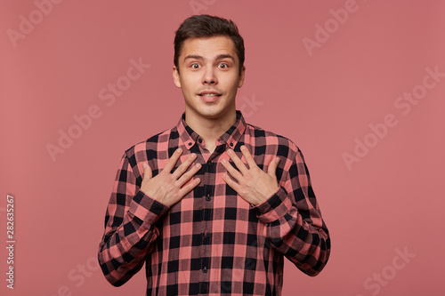 Young attractive wondered man wears in checkered shirt, looks at the camera with happy expression, point at himself, isolated over pink background.