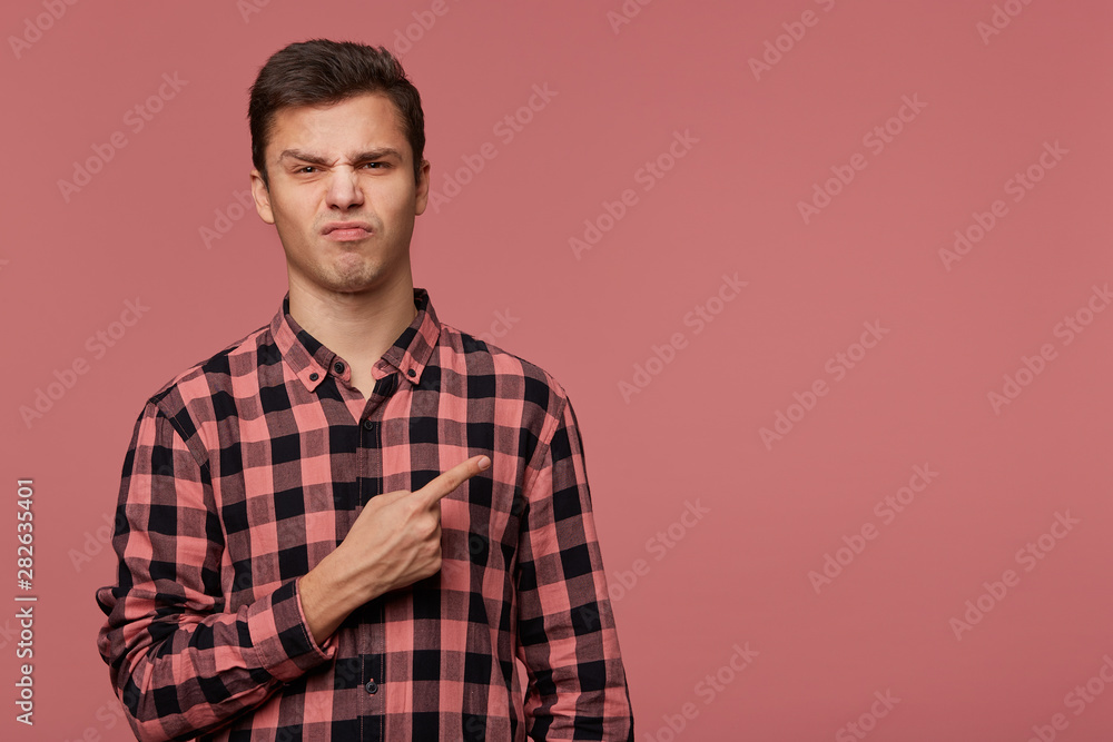 Portrait of frowning cheerful guy wears in checkered shirt, looks at the camera with disgusted expression, wants to turn your attention to copy space at the right side, stands over pink background. Portrait of frowning cheerful guy wears in checkered shirt, looks at the camera with disgusted expression, wants to turn your attention to copy space at the right side, stands over pink background.