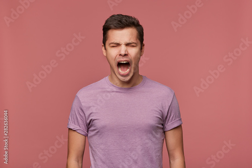 Photo of young attractive guy in blank t-shirt, stands over pink background with closed eyes and screaming, looks angry and unhappy.