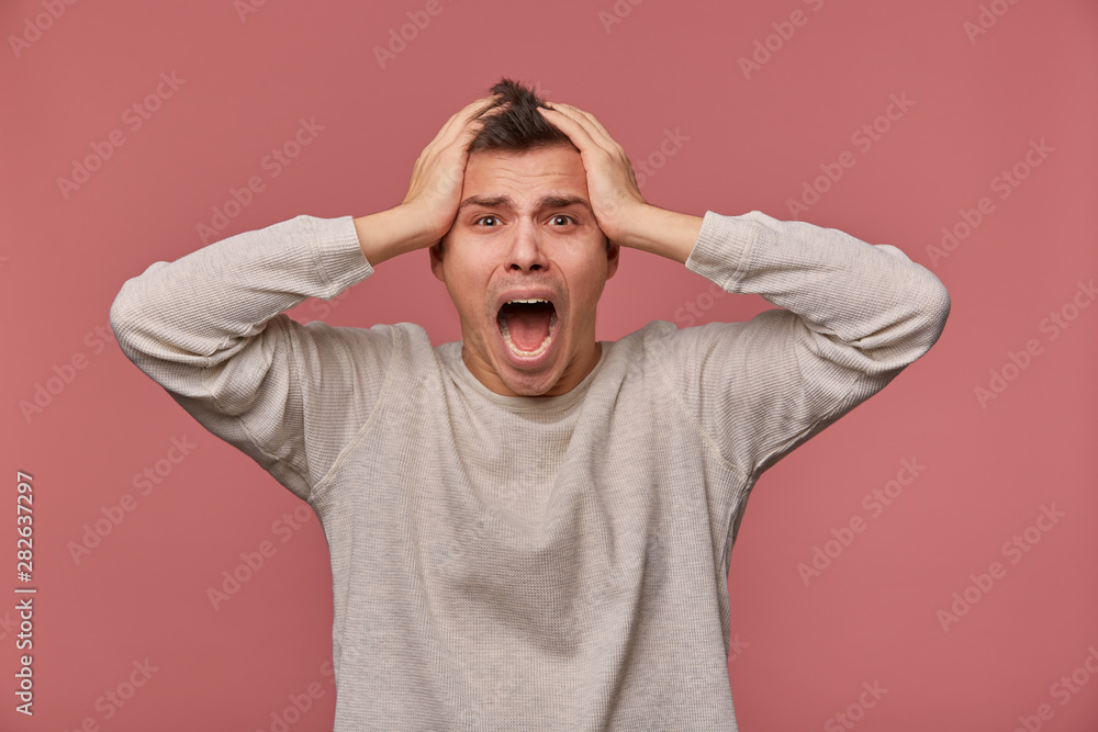 Young scared man in blank long sleeve, holds head and looks at the camera with shocked expression, stands over pink background and screaming, looks afraid. Young scared man in blank long sleeve, holds head and looks at the camera with shocked expression, stands over pink background and screaming, looks afraid.