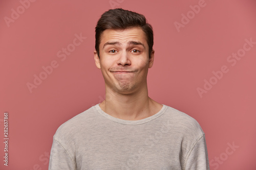 Handsome young sad guy with raised eyebrows wears in blank t-shirt, looks at the camera with mistrust, stands over pink background.