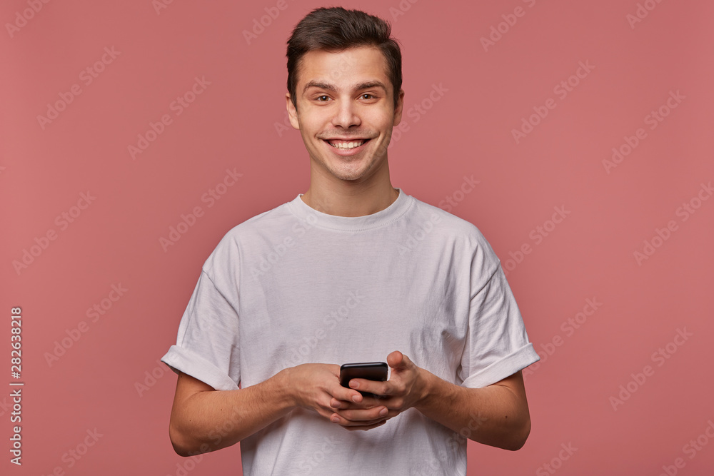 Portrait of young handsome cheerful guy wears in blank t-shirt, looks at the camera with happy expression, stands over pink background and chatting with friend on the telephone. Portrait of young handsome cheerful guy wears in blank t-shirt, looks at the camera with happy expression, stands over pink background and chatting with friend on the telephone.