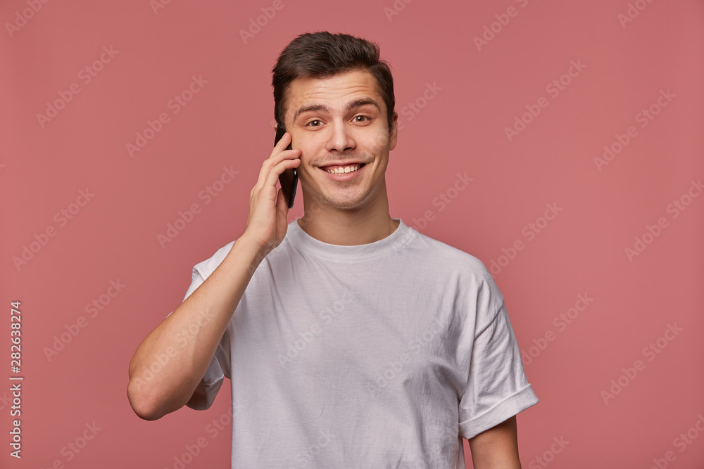 Φωτογραφία Young handsome cheerful man wears in blank t-shirt, looks at the camera with happy expression, stands over pink background and speaks with friend on the telephone