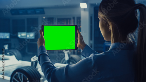 Female Engineer Holds a Tablet Computer with Green Screen Mock Up and Looks at Electric Car Chassis Prototype with Wheels, Batteries and Engine Standing in a High Tech Development Laboratory.