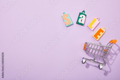 top view of colorful shopping bags with sale inscription falling into toy cart on violet