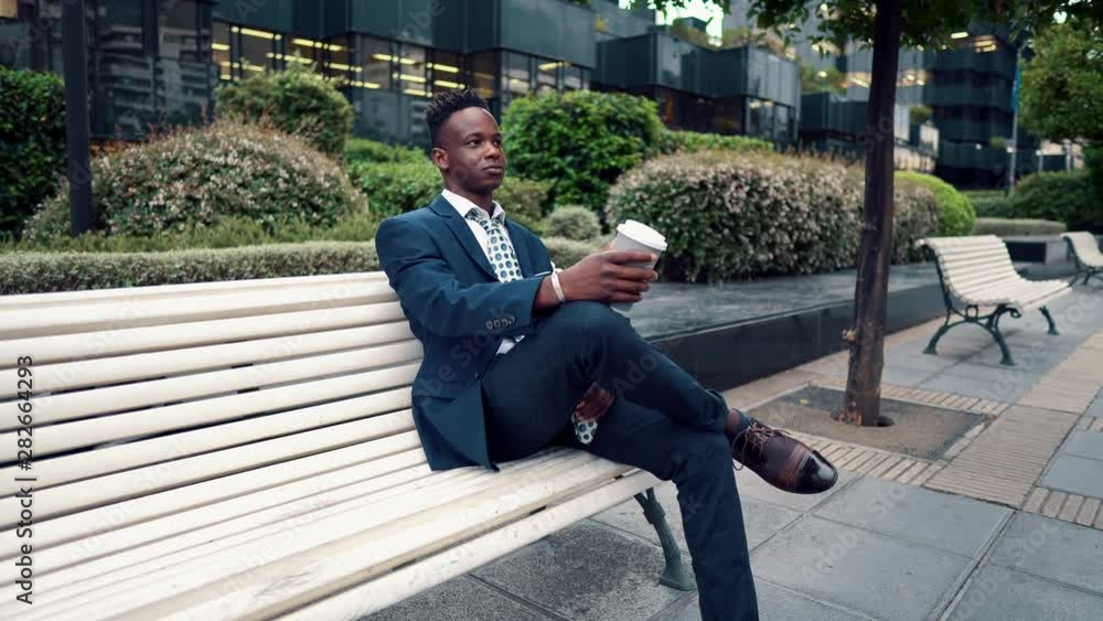 African American businessman wearing blue suit sits on the bench and drinks coffee near office. Business concept African American businessman wearing blue suit sits on the bench and drinks coffee near office. Business concept