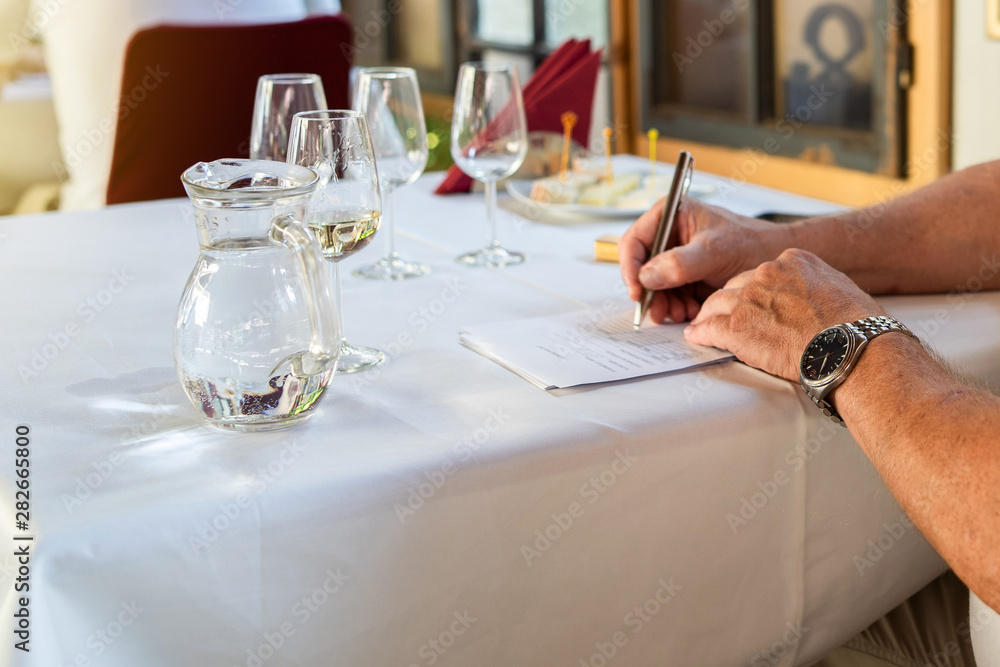close up on hands of Man sommelier expert holding pen with glasses of wine on the table to test it and give marks about the quality to the degustation card