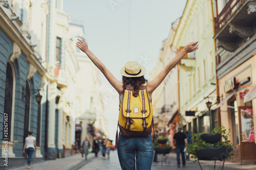 Back view of a young woman traveler with a backpack on her shoulder out sightseeing in a foreign city, stylish female foreigner examines architectural monument during her long-awaited summer vacation