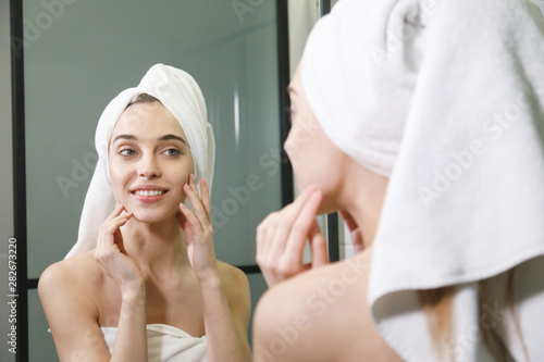 Woman in white bath towel on her head caring of her beautiful skin on the face while standing near the mirror in the bathroom