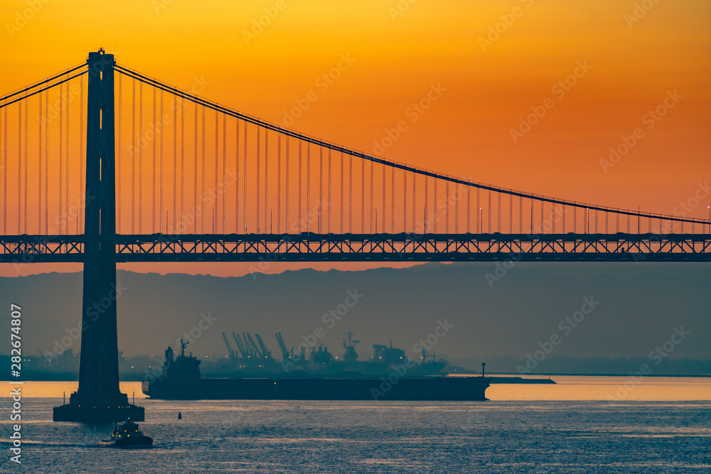 Fototapeta premium Foggy/Misty summer morning sunrise in California and silhouette of the iconic Oakland Bay Bridge in San Francisco as cargo ship sails by. Famous travel location landmark in the west coast city.