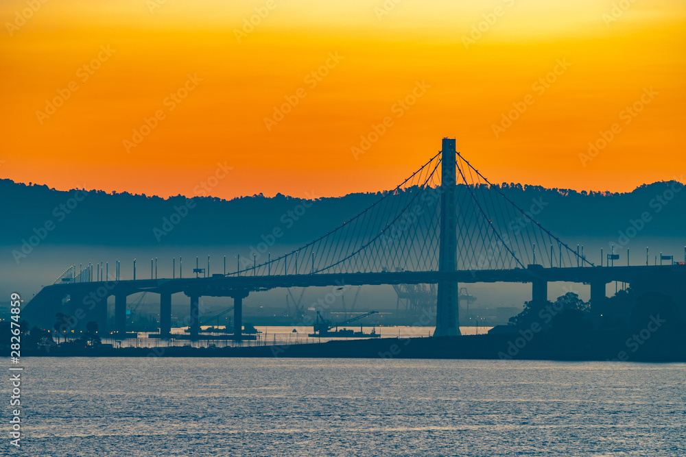 Obraz premium Beautiful summer morning sunrise behind mountain in California gives silhouette to the iconic Oakland Bay Bridge in San Francisco over the ocean.Famous travel location landmark in the west coast city.