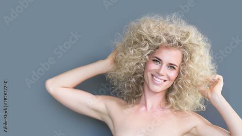 Glad smiling caucasian girl blond curly hairstyle, laughs happily, expresses sincere emotions, admired pleased with new haircut, model in studio with mockup gray or blue background 16:9. Copy space