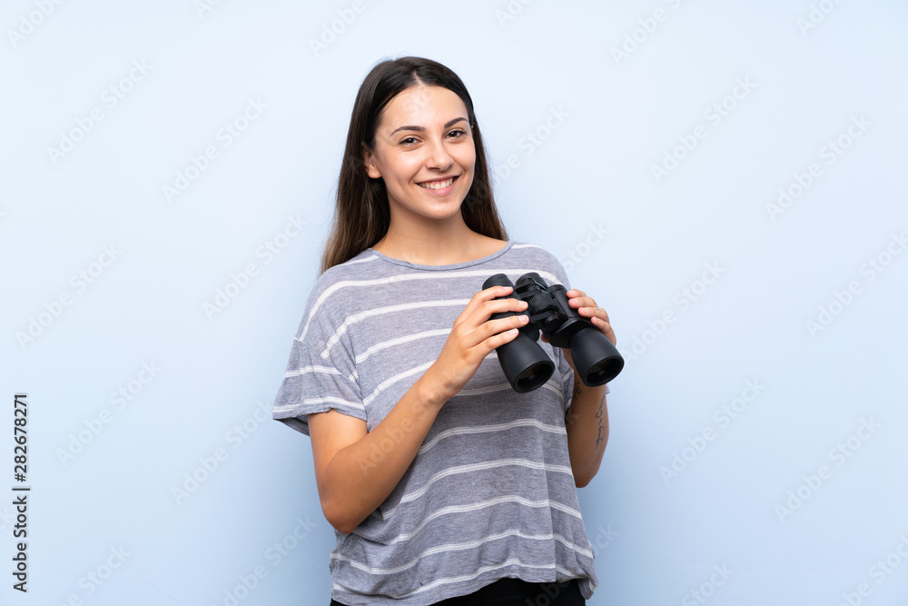 Young brunette woman over isolated blue background with black binoculars