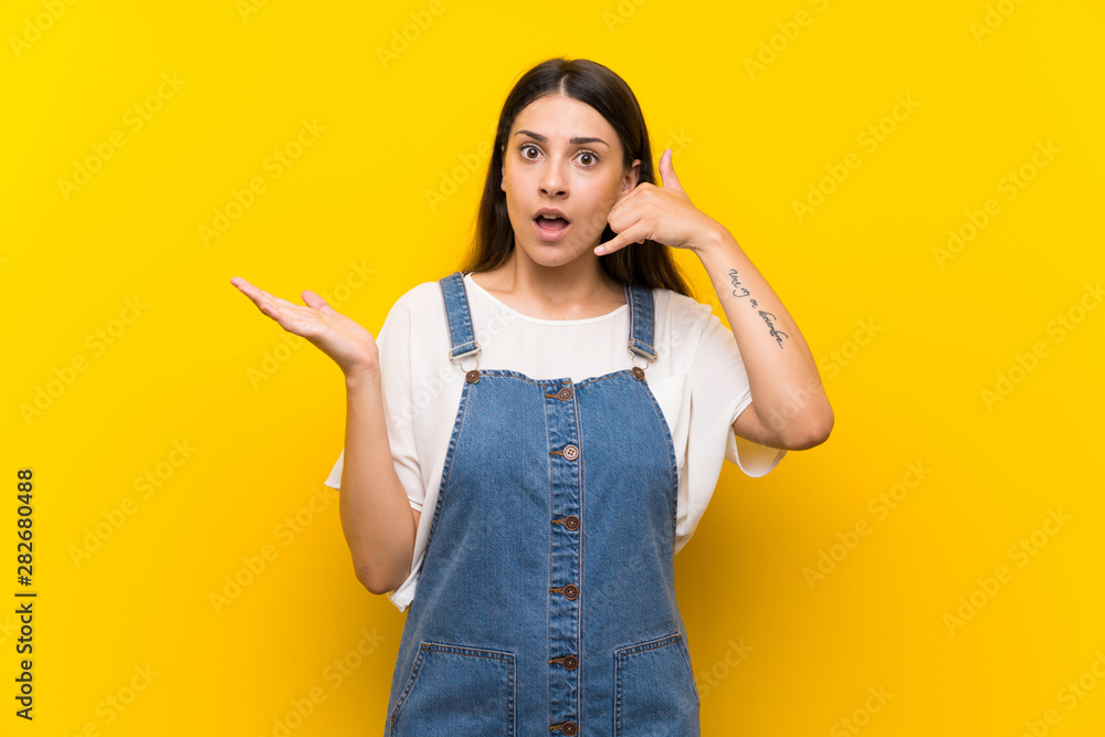 Young woman in dungarees over isolated yellow background making phone gesture and doubting Young woman in dungarees over isolated yellow background making phone gesture and doubting