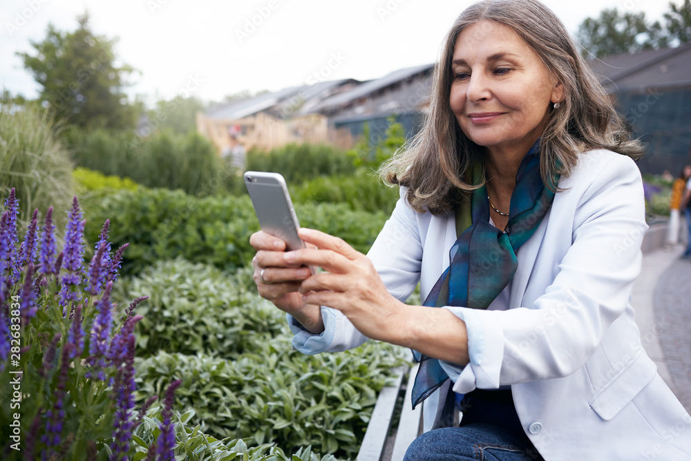 Elegant middle aged European female holding smart phone, trying to capture blossom of violet flowers, walking in park. Smiling mature woman taking picture of beautiful nature around her using mobile Elegant middle aged European female holding smart phone, trying to capture blossom of violet flowers, walking in park. Smiling mature woman taking picture of beautiful nature around her using mobile