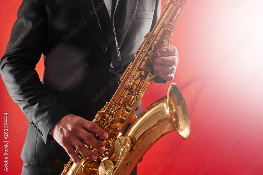 Fototapeta premium Portrait of professional musician saxophonist man in suit plays jazz music on saxophone, red background in a photo studio