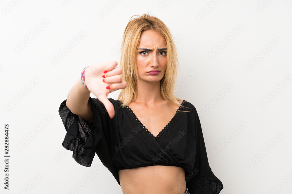 Young blonde woman over isolated white wall showing thumb down with negative expression Young blonde woman over isolated white wall showing thumb down with negative expression