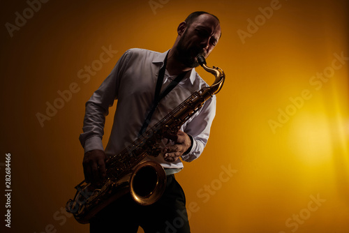 Portrait of professional musician saxophonist man in  white shirt plays jazz music on saxophone, yellow background in a photo studio, side view photo