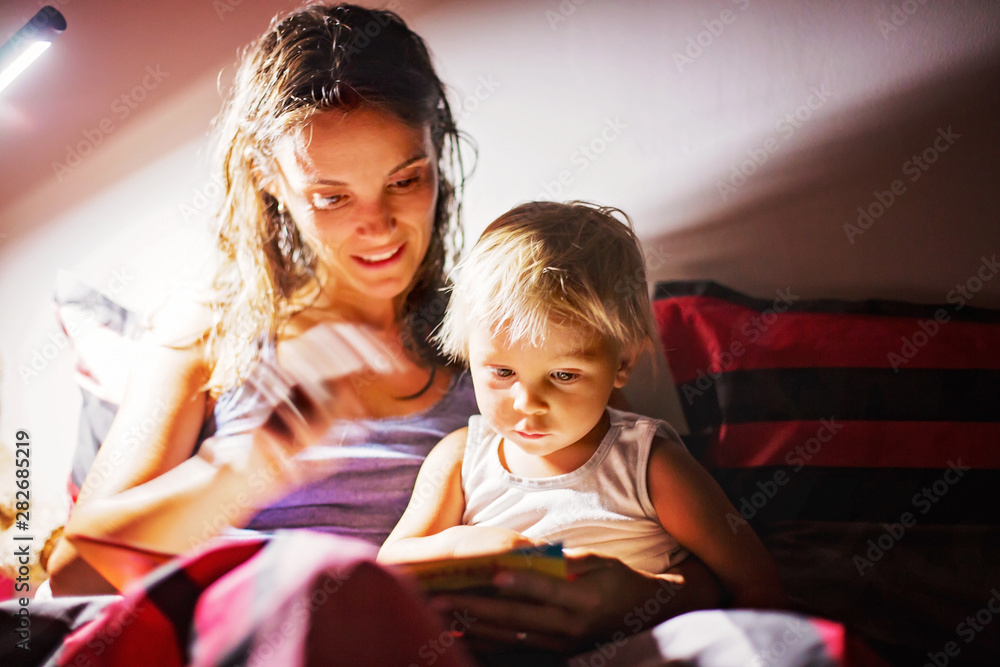 Obraz premium Mother, reading a book to her toddler boy at home at night, night stand lamp turned on