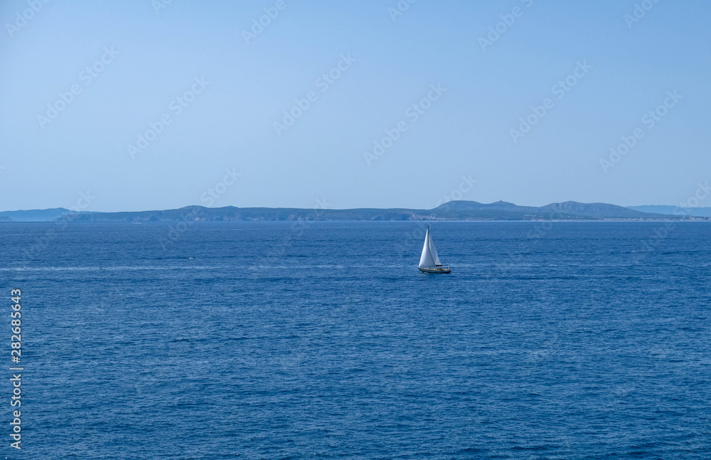 Obraz premium Single white boat sailing on calm water in a sunny day along the coast of the Mediterranean on the background of the islands. Water activities, vacation at sea, happy holidays.