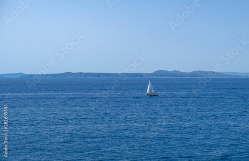 Single white boat sailing on calm water in a sunny day along the coast of the Mediterranean on the background of the islands. Water activities, vacation at sea, happy holidays.