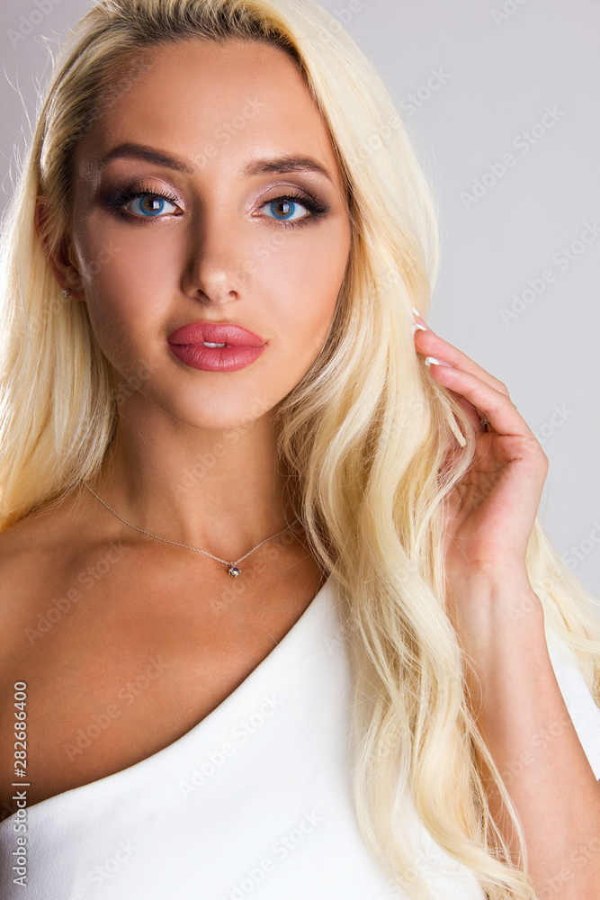 Close-up portrait of sensuality young blonde girl with blue eyes, woman with stylish make up is looking straight and flirty and posing cute with hand near hair on gray wall background, beauty concept Close-up portrait of sensuality young blonde girl with blue eyes, woman with stylish make up is looking straight and flirty and posing cute with hand near hair on gray wall background, beauty concept