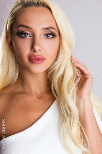 Close-up portrait of sensuality young blonde girl with blue eyes, woman with stylish make up is looking straight and flirty and posing cute with hand near hair on gray wall background, beauty concept