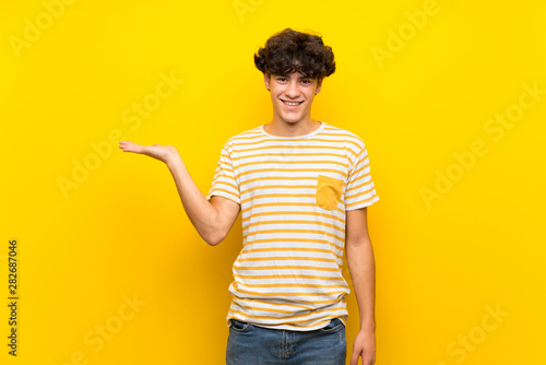 Young man over isolated yellow wall holding copyspace imaginary on the palm to insert an ad
