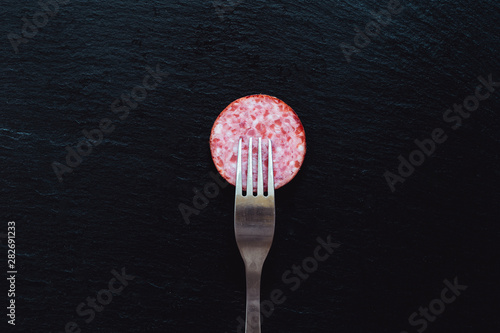 Smoked salami slice with fork on top placed on black background – Spicy traditional homemade meat appetizer on stone textured surface