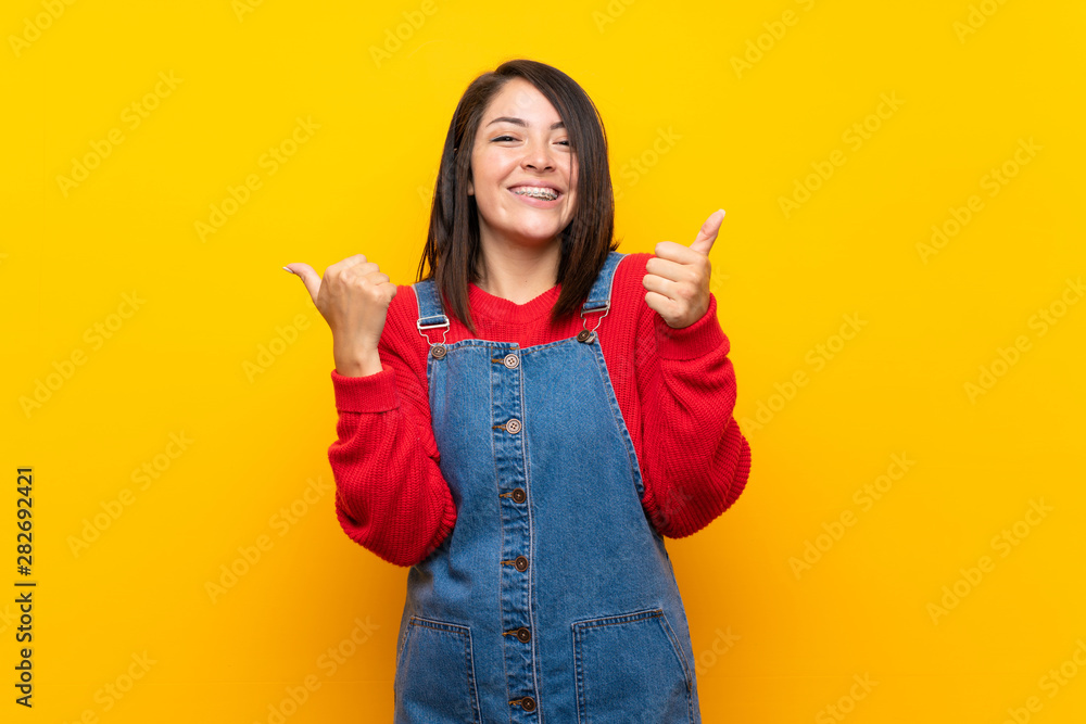 Young Mexican woman with overalls over yellow wall with thumbs up gesture and smiling Young Mexican woman with overalls over yellow wall with thumbs up gesture and smiling